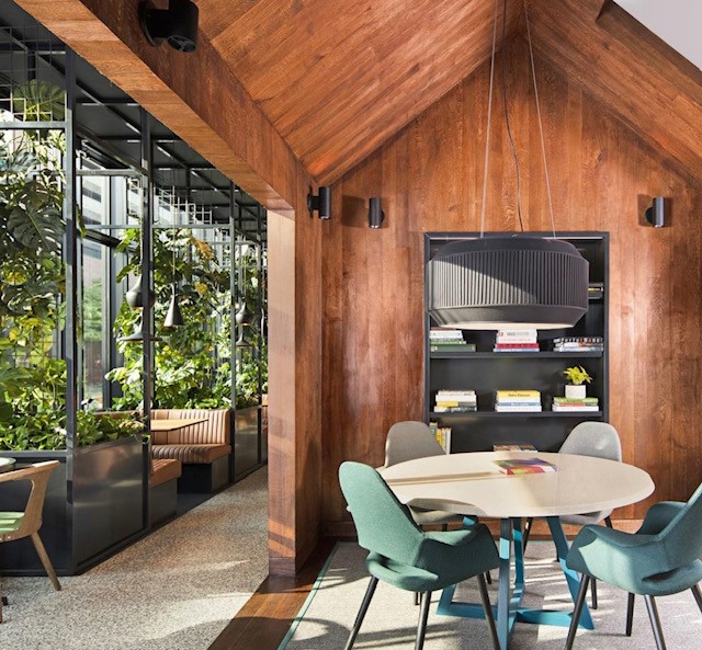 Warm wood-lined café room with a round table, green chairs, plants and pendant light — gathering space at Canard